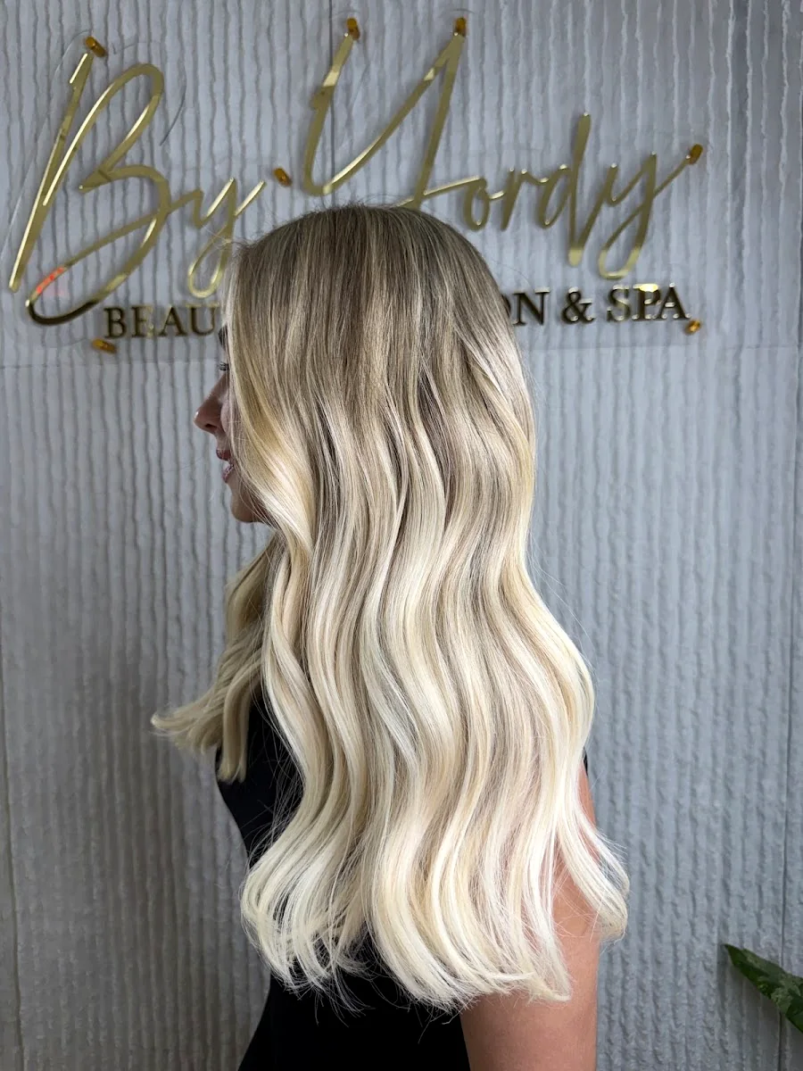 Long sun-kissed balayage styled at By Yordy Beauty Salon & Spa, in front of the salon's gold wall sign